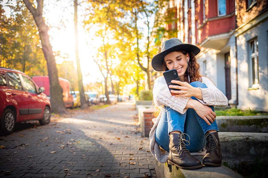 Client Center - Cheerful Woman Sitting on a Ledge in a Residential Neighborhood in the City Using her Phone
