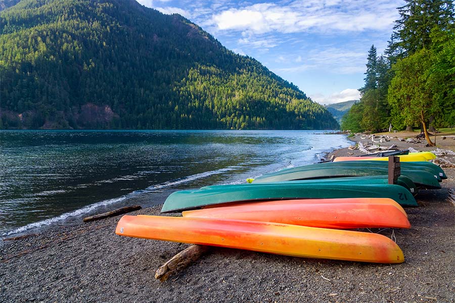 Contact - Colorful Row of Kayaks Resting on the Sand Next to the River with Views of Green Mountains in the Background