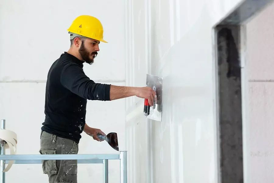 Drywall-Contractor-Insurance-Drywall-Worker-in-Yellow-Helmet-Plastering-Wall-While-Standing-on-Scaffold