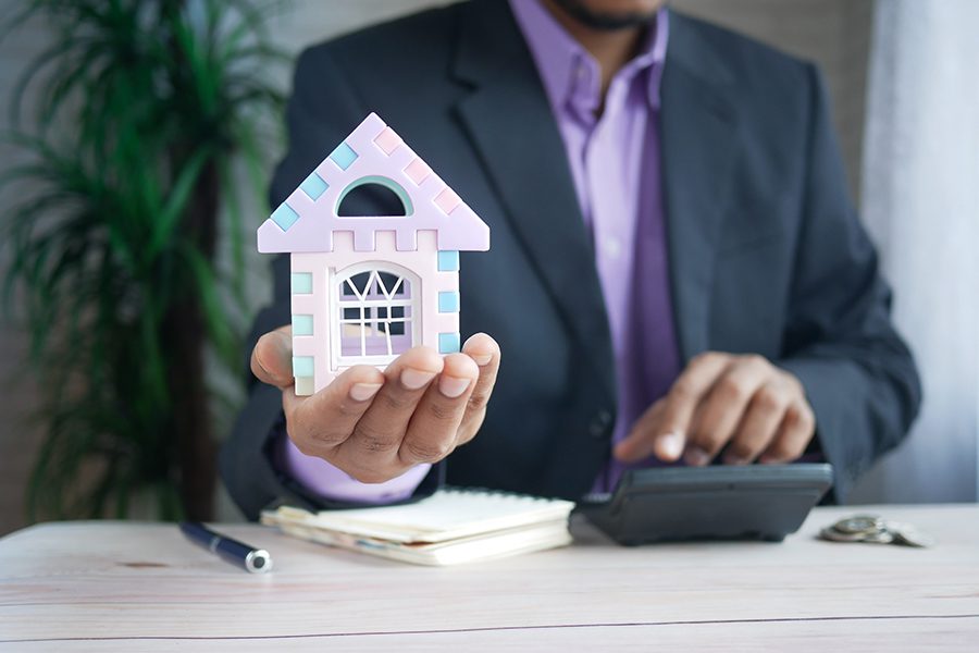 Are You a Mortgage Broker? - Man Holding Out a Plastic Toy Home While He Sits at a Desk Typing on a Calculator Next to a Notepad and Pen